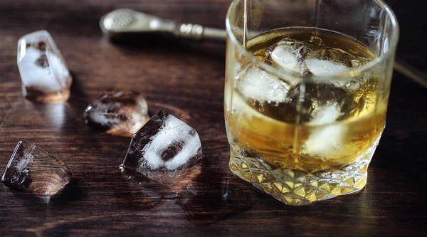 Whiskey with ice with a glass. Cubes of ice on a wooden table and a glass with chilled strong alcohol. Table with American whiskey, bourbon and cards