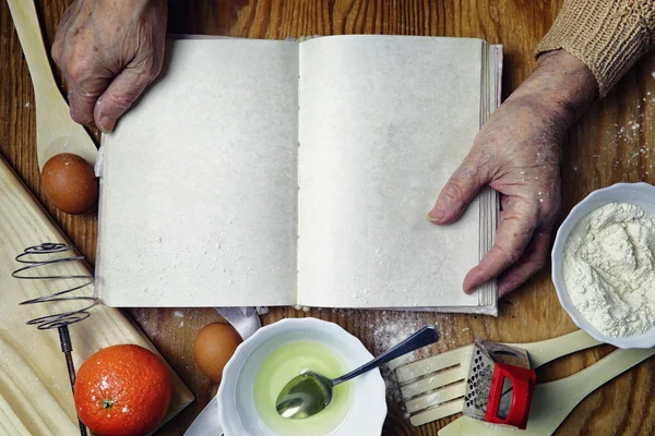 Open recipe book in the hands of an elderly woman in front of a table with utensils