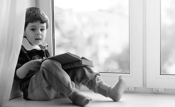 The little boy is reading a book. The child sits at the window a — Stock Photo, Image