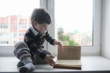 Küçük çocuk bir kitap okuyor. Çocuğun pencerenin önünde oturur bir