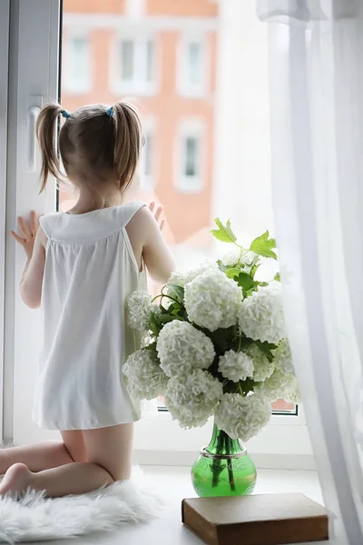 Une petite fille est assise sur le rebord de la fenêtre. Un bouquet de fleurs — Photo