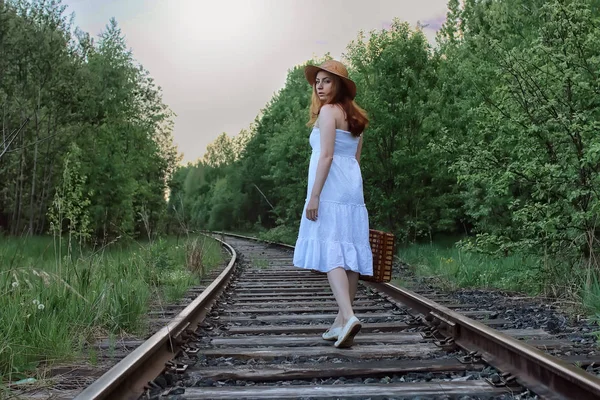 Beyaz elbiseli ve hasır bavullu bir kız raylarda yürüyor. — Stok fotoğraf