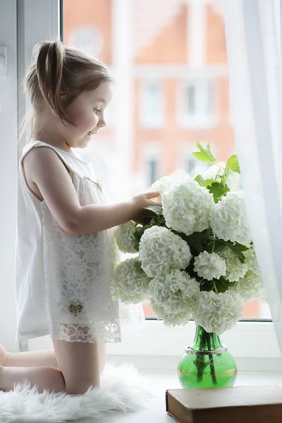 Une petite fille est assise sur le rebord de la fenêtre. Un bouquet de fleurs — Photo