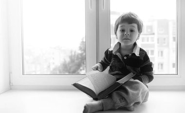 Küçük çocuk bir kitap okuyor. Çocuğun pencerenin önünde oturur bir