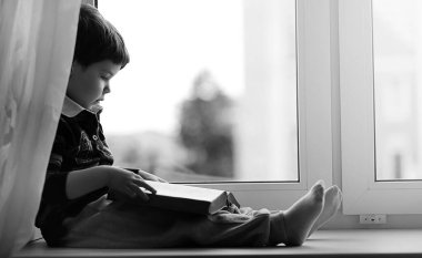 Küçük çocuk bir kitap okuyor. Çocuğun pencerenin önünde oturur bir