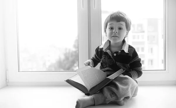 Het jongetje is het lezen van een boek. Het kind zit bij het raam een — Stockfoto