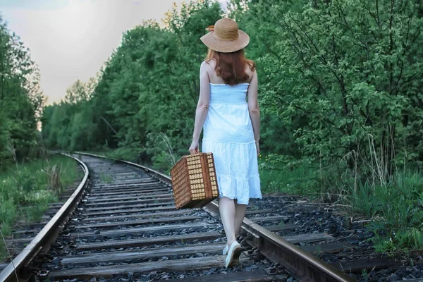 Menina em um vestido de verão branco e mala de vime andando sobre trilhos — Fotografia de Stock