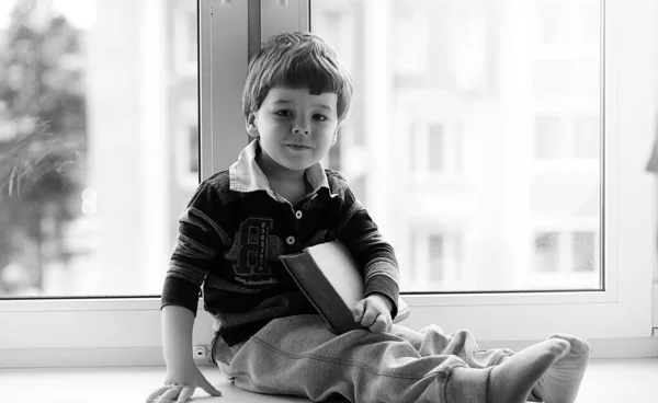 Het jongetje is het lezen van een boek. Het kind zit bij het raam een — Stockfoto