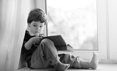 Küçük çocuk bir kitap okuyor. Çocuğun pencerenin önünde oturur bir