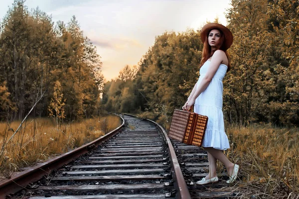 Güz parkındaki beyaz elbiseli kız ve hasır bir bavul yürüyor.