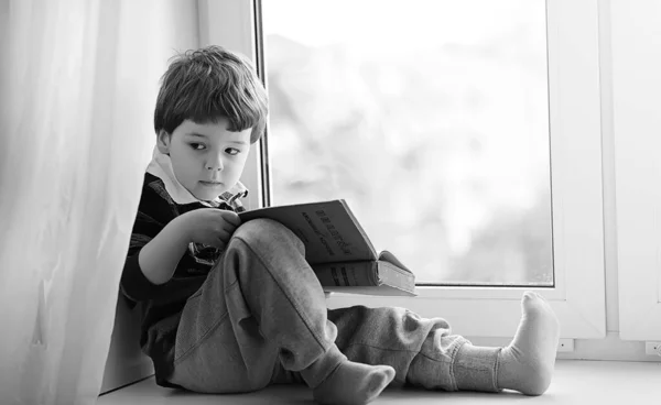Küçük çocuk bir kitap okuyor. Çocuğun pencerenin önünde oturur bir — Stok fotoğraf