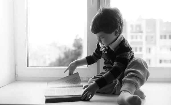 Den lilla pojken läser en bok. Barnet sitter vid fönstret en — Stockfoto