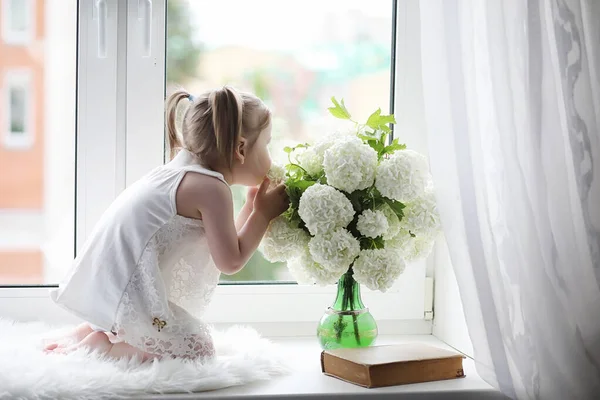 Une petite fille est assise sur le rebord de la fenêtre. Un bouquet de fleurs — Photo