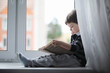 Küçük çocuk bir kitap okuyor. Çocuğun pencerenin önünde oturur bir