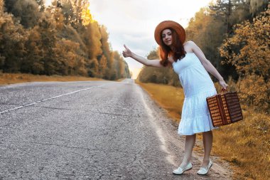 Güz parkındaki beyaz elbiseli kız ve hasır bir bavul yürüyor.