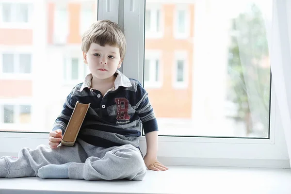 Küçük çocuk bir kitap okuyor. Çocuğun pencerenin önünde oturur bir — Stok fotoğraf