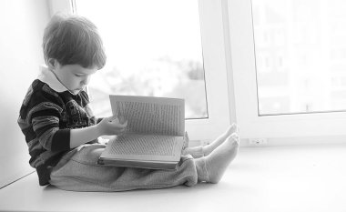 Küçük çocuk bir kitap okuyor. Çocuğun pencerenin önünde oturur bir