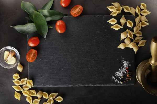 Cáscaras de pasta sobre piedra negra. Tomates y hojas de laurel con allspi — Foto de Stock