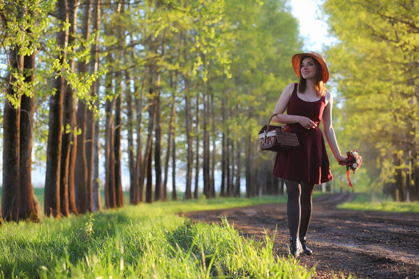 A girl in a hat on a walk in the park. A girl with a basket walks in the spring. Girl is walking along the road at sunset
