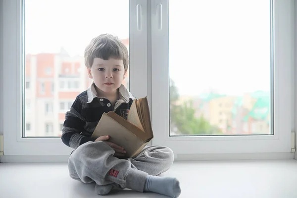 Küçük çocuk bir kitap okuyor. Çocuğun pencerenin önünde oturur bir — Stok fotoğraf