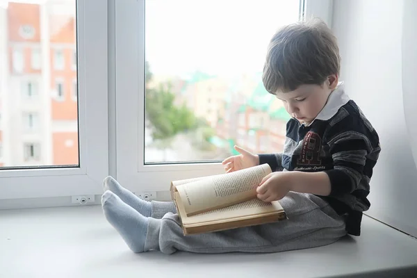 Küçük çocuk bir kitap okuyor. Çocuğun pencerenin önünde oturur bir — Stok fotoğraf