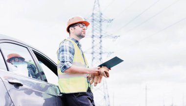 Kasklı ve üniformalı bir adam, sahada bir elektrikçi. Profesyonel elektrik mühendisi elektrik hatlarını inceliyor..