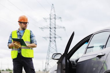 Kasklı ve üniformalı bir adam, sahada bir elektrikçi. Profesyonel elektrik mühendisi elektrik hatlarını inceliyor..