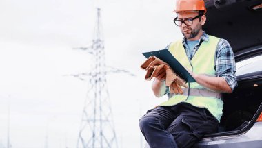 Kasklı ve üniformalı bir adam, sahada bir elektrikçi. Profesyonel elektrik mühendisi elektrik hatlarını inceliyor..