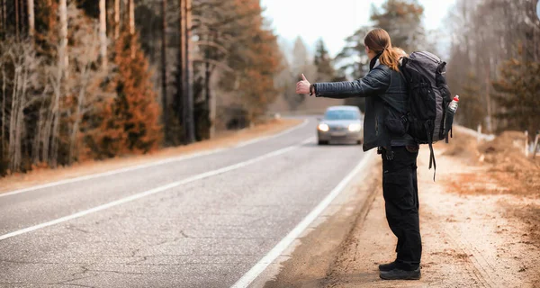 田舎では若い男がヒッチハイクをしている 男は旅行のために通りすがりの車を捕まえようとしている リュックを持った男が南へヒッチハイクに行った — ストック写真