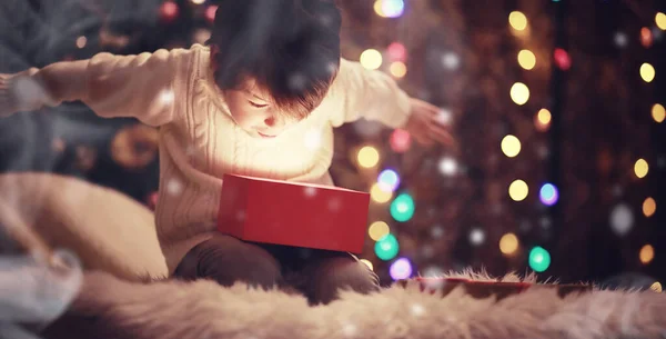 Family on Christmas eve at fireplace. Little boy opening Xmas presents. Child under Christmas tree with gift boxes. Decorated living room with traditional fire place. Cozy warm winter evening home.