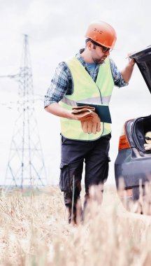 Kasklı ve üniformalı bir adam, sahada bir elektrikçi. Profesyonel elektrik mühendisi elektrik hatlarını inceliyor..