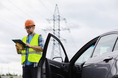 Kasklı ve üniformalı bir adam, sahada bir elektrikçi. Profesyonel elektrik mühendisi elektrik hatlarını inceliyor..
