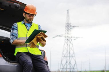 Kasklı ve üniformalı bir adam, sahada bir elektrikçi. Profesyonel elektrik mühendisi elektrik hatlarını inceliyor..