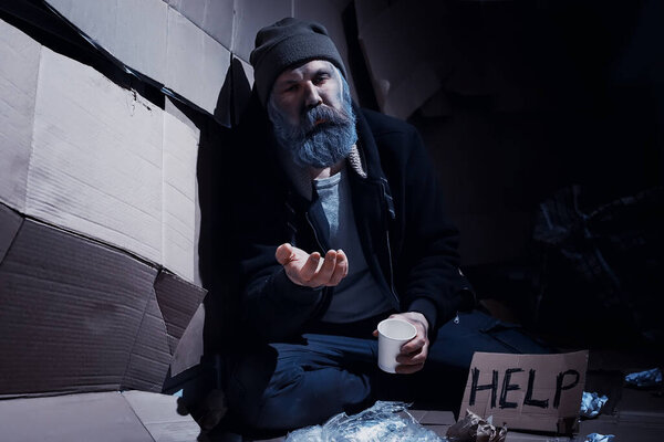 A homeless bearded man sits on boxes on street and asks for help. Need a homeless person asks for money for food and overnight.