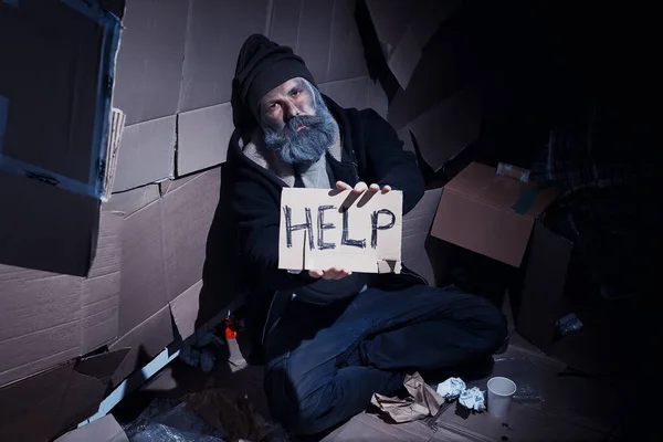 ホームレスの髭を生やした男が通りの箱の上に座って助けを求める ホームレスの人が食料と一夜のためにお金を要求する必要がある — ストック写真