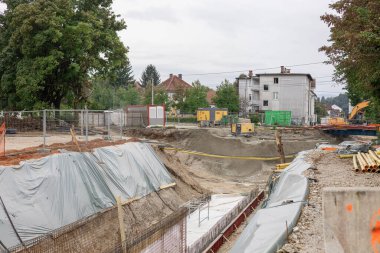 Ika, Ljubljana 'daki kentsel kazı alanı: Gri muşambalarla kaplı eğimli toprak bankalarının ortasındaki derin beton kaplı siper. Turuncu güvenlik çitleri toprak yığınlarını, kamyonları, kazıcıları ve bulutlu gökyüzünün altında dağlarla çevrili uzak binaları sarıyor..