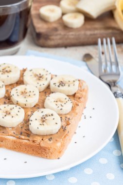 Sağlıklı kahvaltı sandviçi, fıstık ezmeli, muz ve chia tohumlu, beyaz tabakta, dikey.