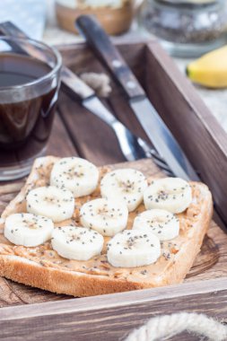 Sağlıklı sandviç, fıstık ezmeli, muzlu ve chia tohumlu, bir tepside, bir fincan kahveyle birlikte, dikey