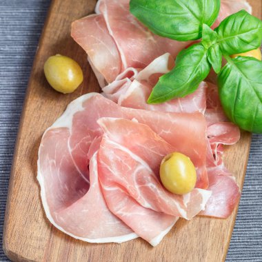 Ahşap bir tahtada jambon, fesleğen yaprakları ve arka planda yeşil zeytinler, kare şeklinde.