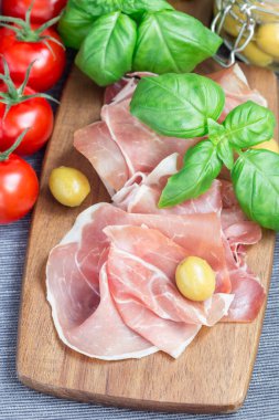 Ahşap bir tahtada jambon, yeşil zeytin, domates ve fesleğen yapraklı, dikey.