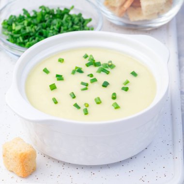 Patates püresi çorbası beyaz seramik kasede yeşil soğanla süslenmiş, kızarmış ekmeklerle servis edilmiş, kare şeklinde, yakın plan.