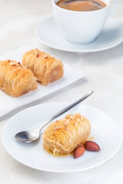 Yunan pastası Kataifi, dilimlenmiş fileto hamuru, badem fıstığıyla doldurulmuş, bal şurubu, beyaz bir tabakta, kahve ile servis edilmiş, dikey.