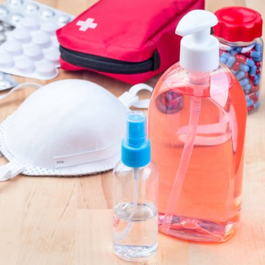 İlk yardım çantasında haplar, maske, kızılötesi vücut termometresi, dezenfektan, ahşap masada farklı ilaçlar, kare format.