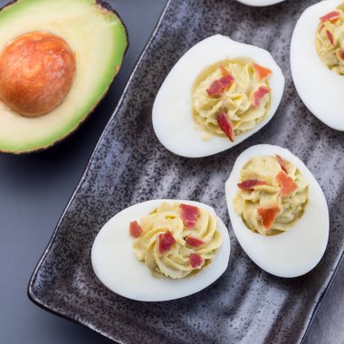 Avokado, yumurta sarısı ve mayonez ile doldurulmuş yumurtalar, pastırmayla süslenmiş, üst manzara, kare format.