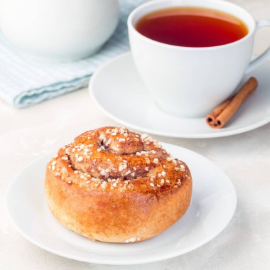 Beyaz tabakta bir tarçınlı çörek, bir fincan kırmızı çay, kare formatta, yakın çekim.