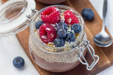 Kavanozda katmanlı çikolata ve fıstık ezmeli chia tohumu pudingi, ahududu, yabanmersini, bal ve hindistan cevizi taneleri ile süslenmiş, yatay, yakın plan