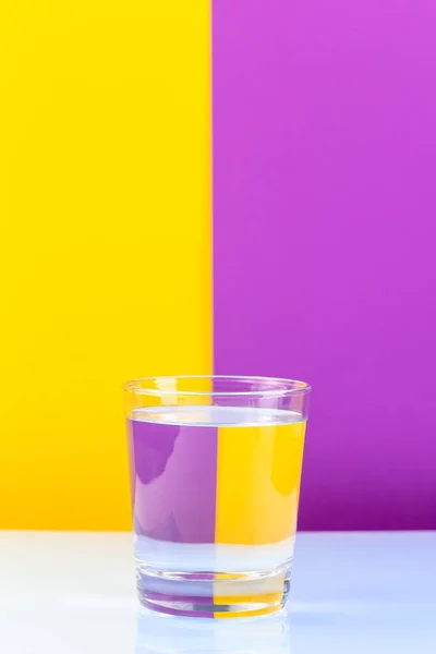 Złudzenie Optyczne Szklanką Wody Kolorowym Papierem Żółte Fioletowe Tło Pionowe — Zdjęcie stockowe