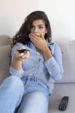 Kanepede oturan korkmuş genç bir kadın uzaktan kumandayla televizyon izliyor, korku filmi seyrediyor.