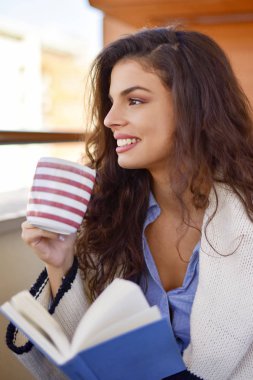 Genç bir kadın kitap okuyor ve balkonda çay içiyor.