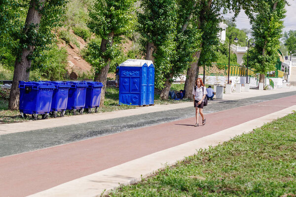 Оренбург, Россия - 1 июня 2019 года: Набережная Урала. Саммер. Девушка идет по тропинке мимо мусорных баков, сухих шкафов и мигрантов, отдыхающих в тени деревьев
